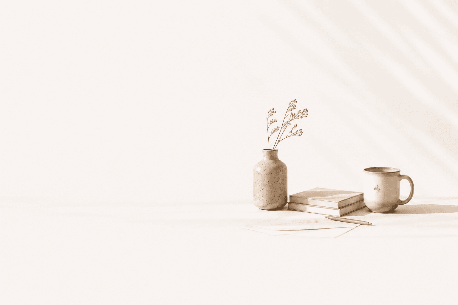 A speckled ceramic vase of dried flowers beside two stacked books, a slim pen and a pale mug, on linen in soft morning light.